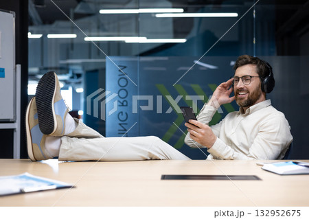 Relaxed and smiling young man sitting in the office wearing headphones, legs crossed on the desk, listening to music from his mobile phone. Relaxed and smiling young man sitting in the office wearing headphones, legs crossed on the desk, listening to music from his mobile phone. 132952675