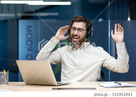 A relaxed young man in headphones sits in the office at a table with a laptop and listens to music while dancing. 132952681