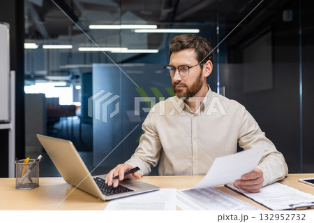 A concentrated young man is sitting in the office at a desk, holding documents in his hands and working on a laptop. 132952732
