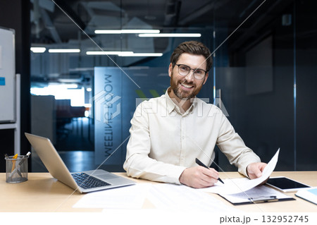 Portrait of a smiling young man working in the office. Sitting at a desk, working on a laptop and with documents. Portrait of a smiling young man working in the office. Sitting at a desk, working on a laptop and with documents. 132952745