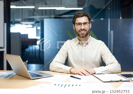 Portrait of a young smiling man in a shirt and glasses sitting in the office at a table with a laptop and documents, looking at the camera. Portrait of a young smiling man in a shirt and glasses sitting in the office at a table with a laptop and documents, looking at the camera. 132952755
