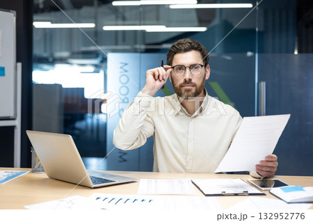 Portrait of a young man working in an office with documents, sitting at a table, supporting his glasses with his hand, looking at the camera. Portrait of a young man working in an office with documents, sitting at a table, supporting his glasses with his hand, looking at the camera. 132952776