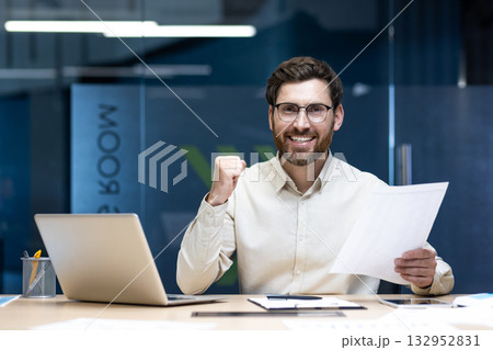Portrait of a happy young male office worker sitting at his desk, holding documents in his hand and showing a victory sign at the camera. Portrait of a happy young male office worker sitting at his desk, holding documents in his hand and showing a victory sign at the camera. 132952831