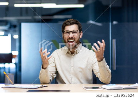 Close-up portrait of an angry and irritated young man sitting in an office at a desk and yelling at the camera while waving his hands. Close-up portrait of an angry and irritated young man sitting in an office at a desk and yelling at the camera while waving his hands. 132953624