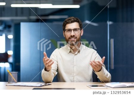 Portrait of a young man sitting in the office at a table in front of the camera and talking on a video call, gesturing to the camera. 132953654