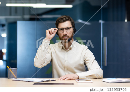 Portrait of a serious young man sitting in front of the camera in a headset in the office at the desk, looking at the camera and holding glasses. Portrait of a serious young man sitting in front of the camera in a headset in the office at the desk, looking at the camera and holding glasses. 132953793