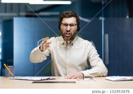Portrait of an angry young man in a headset sitting in the office at a desk and talking angrily and pointing his finger at the camera. 132953876