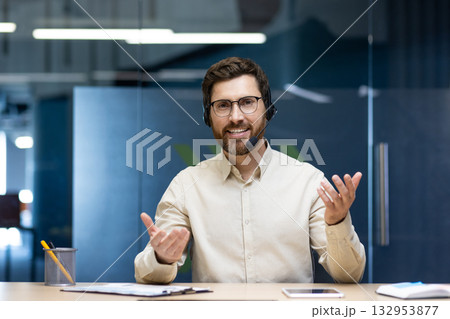 A smiling young man wearing a headset is sitting at a desk in the office and talking to the camera remotely gesturing with his hands. 132953877