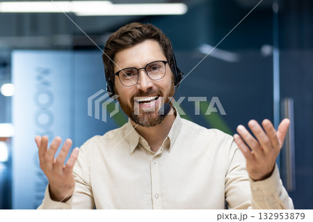 Close-up portrait of a young man wearing a headset and shirt sitting in the office at a desk, smiling and talking to the camera and gesturing with his hands. Close-up portrait of a young man wearing a headset and shirt sitting in the office at a desk, smiling and talking to the camera and gesturing with his hands. 132953879