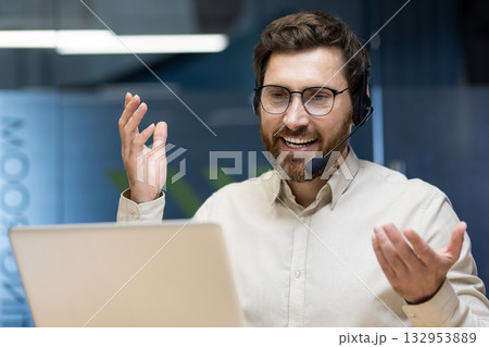 Close-up photo of a young smiling man in a headset sitting in the office at a table in front of a laptop and talking on a video call. Close-up photo of a young smiling man in a headset sitting in the office at a table in front of a laptop and talking on a video call. 132953889