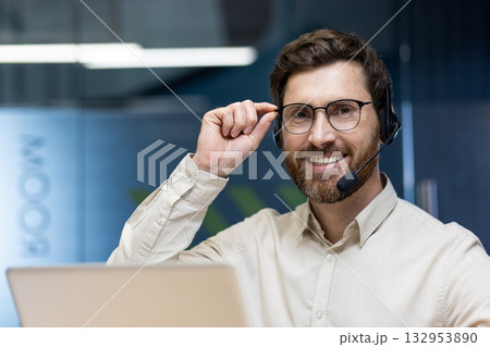 Close-up photo of a young man in a headset sitting at a desk in the front office, working on a laptop, smiling and looking at the camera while holding his glasses. Close-up photo of a young man in a headset sitting at a desk in the front office, working on a laptop, smiling and looking at the camera while holding his glasses. 132953890
