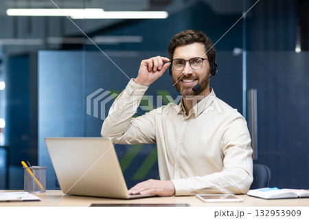 Portrait of a young smiling man wearing a headset sitting at a desk in the office, working on a laptop, holding glasses and looking at the camera. 132953909