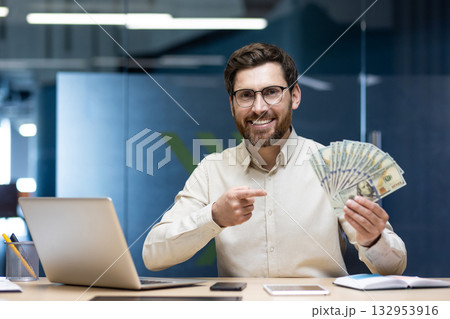 Portrait of a smiling young man sitting in an office at a desk with a laptop, holding a fan of banknotes and pointing at dollars. 132953916