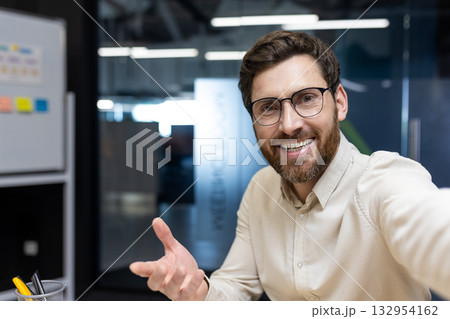 Close-up photo of a young smiling man in a shirt and glasses who is in the office, holding a camera in his hands and talking on the Internet, gesturing to the camera with his hand. Close-up photo of a young smiling man in a shirt and glasses who is in the office, holding a camera in his hands and talking on the Internet, gesturing to the camera with his hand. 132954162