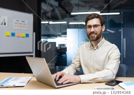 Portrait of a smiling young man sitting in the office behind a laptop and looking smugly at the camera. Portrait of a smiling young man sitting in the office behind a laptop and looking smugly at the camera. 132954168