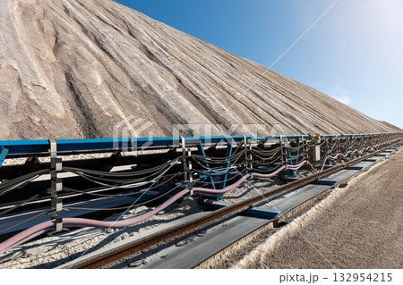 Long blue conveyor belt transports white potash ore material toward massive potassium mining dump sunlight. Industrial machinery stretches distance mineral extraction continuous production process 132954215