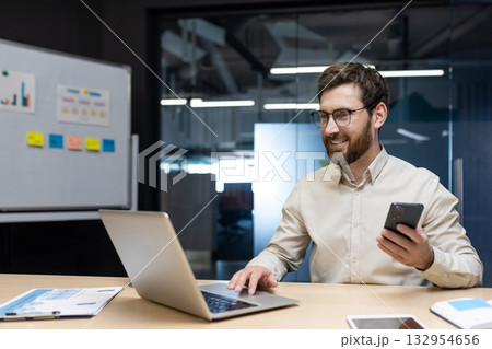 Smiling young man businessman, developer works in office at laptop on whiteboard with stickers, types on keyboard, holds mobile phone in hand. 132954656