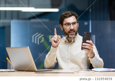 A young businessman is sitting in the office at a desk with a laptop, talking on a video call on a mobile phone camera, pointing his index finger at the screen. 132954659