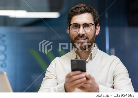 Close-up portrait of a young smiling businessman in glasses and a shirt who is in the office, holding a mobile phone and looking at the camera. Close-up portrait of a young smiling businessman in glasses and a shirt who is in the office, holding a mobile phone and looking at the camera. 132954665