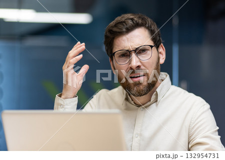 Close-up portrait of an angry and frustrated young male office worker sitting at a workplace with a laptop and looking annoyed at the camera while gesturing with his hand. 132954731