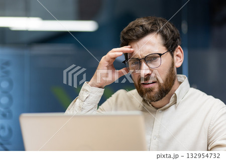 Close-up portrait of a young upset male businessman sitting in the office in front of a laptop and holding his head in frustration. 132954732