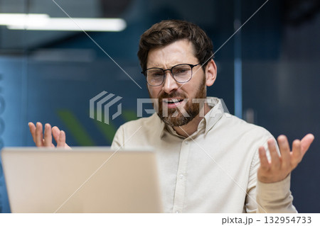 Angry young man sitting in the office with a desk, looking at the laptop screen and throwing up his hands in frustration. 132954733