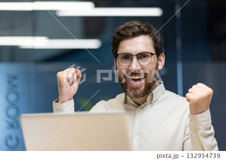 A close-up portrait of a young happy and successful businessman who is in the office, sitting at the workplace and is happy to the camera, showing a victory gesture with his hands. 132954739