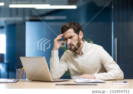 Confident young man sitting in the office at the desk, holding his head and looking worriedly at the laptop. Confident young man sitting in the office at the desk, holding his head and looking worriedly at the laptop. 132954751
