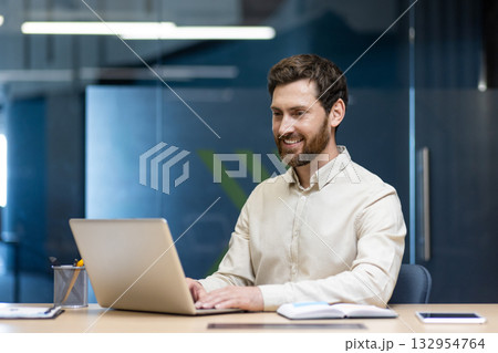 A young male businessman works in the office at a laptop, sits at a desk and looks at the screen with a smile. A young male businessman works in the office at a laptop, sits at a desk and looks at the screen with a smile. 132954764