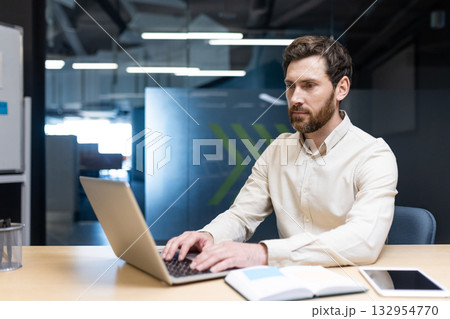 A serious young man in a light-colored shirt is sitting in the office at a desk and is concentrating on working on a laptop. A serious young man in a light-colored shirt is sitting in the office at a desk and is concentrating on working on a laptop. 132954770