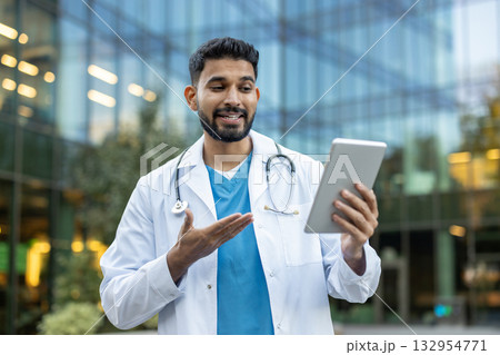 Indian doctor wearing a lab coat and stethoscope is having a video consultation on a digital tablet, discussing healthcare services and modern medical technology outdoors 132954771