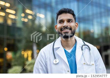 Confident medical professional wearing white coat and stethoscope smiles while looking away from the camera and standing outside modern hospital building 132954777