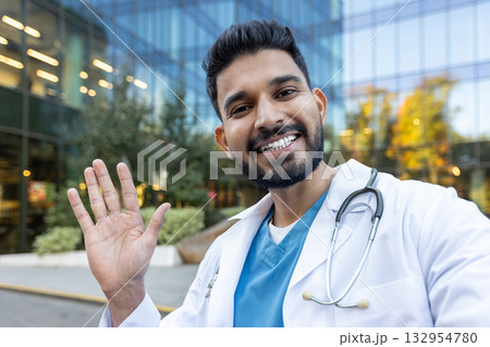Portrait of a smiling indian doctor waving hand in front of a modern hospital building, radiating friendliness and approachability while showcasing confidence and professionalism Portrait of a smiling indian doctor waving hand in front of a modern hospital building, radiating friendliness and approachability while showcasing confidence and professionalism 132954780