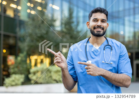 Young male doctor wearing blue scrubs and stethoscope is pointing with both index fingers to different directions in front of a modern hospital building, smiling and looking at camera Young male doctor wearing blue scrubs and stethoscope is pointing with both index fingers to different directions in front of a modern hospital building, smiling and looking at camera 132954789