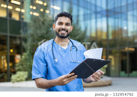 Confident and friendly indian doctor is standing outside a modern hospital building, holding a clipboard with patient medical records and smiling 132954792