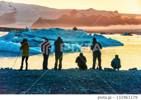 Tourists admire the stunning sunset over Iceland's Glacier Lagoon, surrounded by large icebergs. The serene scene captures the beauty of nature 132961179