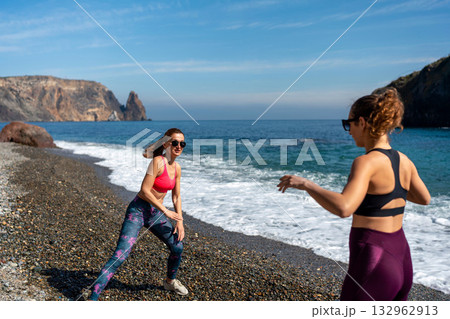 Fitness women stretching routine on pebble beach by scenic sea cliffs 132962913