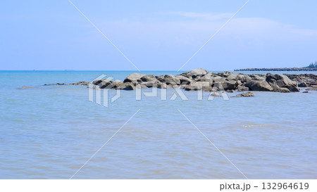 Rocky Breakwater on a Tropical Seashore Against a Bright Blue Sky and Calm Blue Ocean Water 132964619