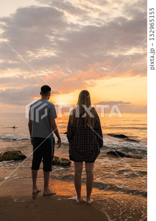 Couple standing barefoot on the beach and watching the sunrise over the calm sea with golden reflections on the water Couple standing barefoot on the beach and watching the sunrise over the calm sea with golden reflections on the water 132965155