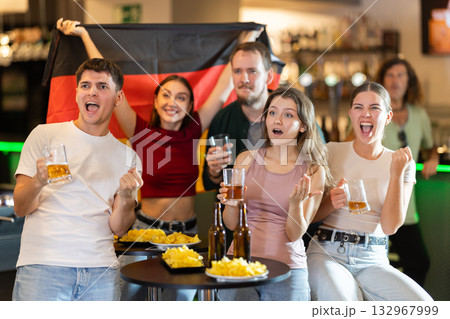 Group of fans of Germany team with a flag are watching the match on TV in beer bar. They are worried and supporting their 132967999