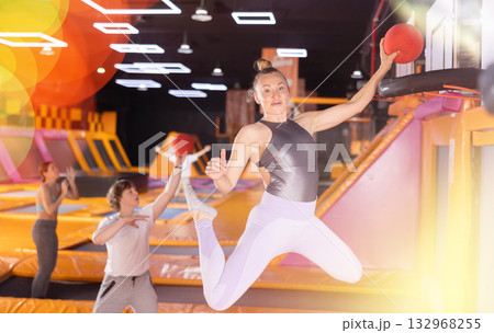 Energized woman enjoying amusement activities while jumping high on trampoline and hitting basketball hoop with ball in game club Energized woman enjoying amusement activities while jumping high on trampoline and hitting basketball hoop with ball in game club 132968255