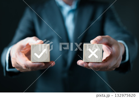 A businessman in a suit presents two wooden blocks, one with a white checkmark and the other with a white X, symbolizing decision making, evaluation, approval, rejection, and business choices. A businessman in a suit presents two wooden blocks, one with a white checkmark and the other with a white X, symbolizing decision making, evaluation, approval, rejection, and business choices. 132975620