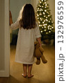Little girl standing in doorway holding teddy bear looking at christmas tree 132976559