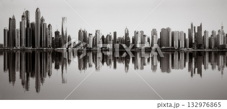 Monochrome city skyline with tall skyscrapers reflecting on calm water under a cloudy sky, Dubai Marina bay UAE 132976865
