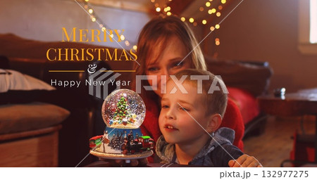 Mother and son wearing red sweater, hoodie holding snow globe in living room, with fairy lights 132977275