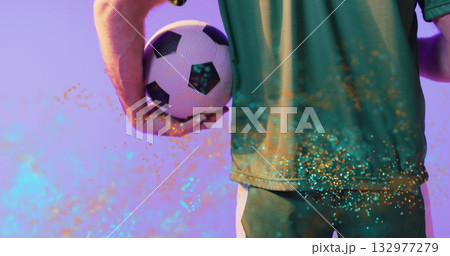Athlete wearing green jersey and shorts holding soccer ball behind waist in studio, with confetti 132977279