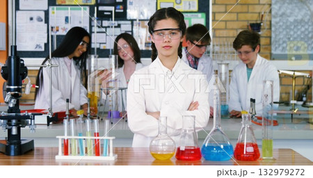 Girl wearing lab coat and goggles overseeing classmates mixing chemicals in school lab, with flasks 132979272