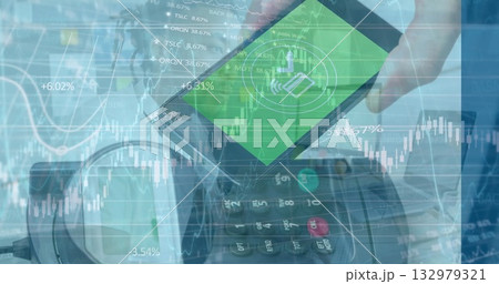 Holding smartphone showing payment icon hovering over card reader at checkout with financial charts 132979321