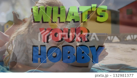 Lying girl focusing on tablet at sandy beach, with striped towel and colorful wooden huts 132981377