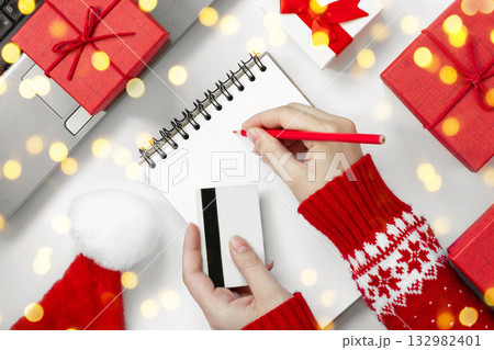 Female Hands with Credit Card and Pencil on a Desk. Christmas Online Shopping Concept. Girl Writing Wish List. Christmas Sweater. Laptop, Red Gift Boxes, Notepad on White Table. Winter Holidays Sale. 132982401